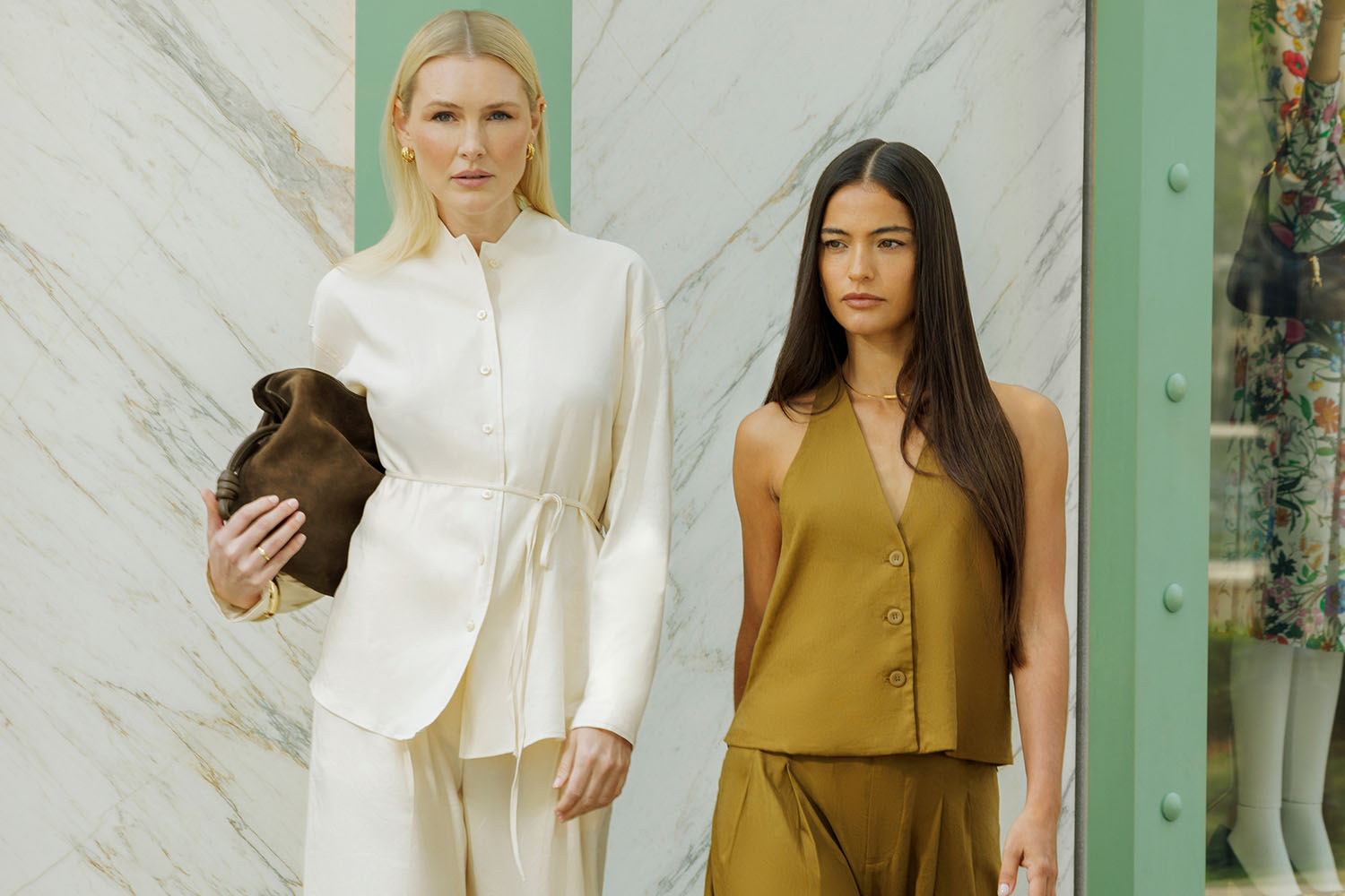 Two women in cream and olive outfits standing outside a boutique with marble walls and mint-green trim.